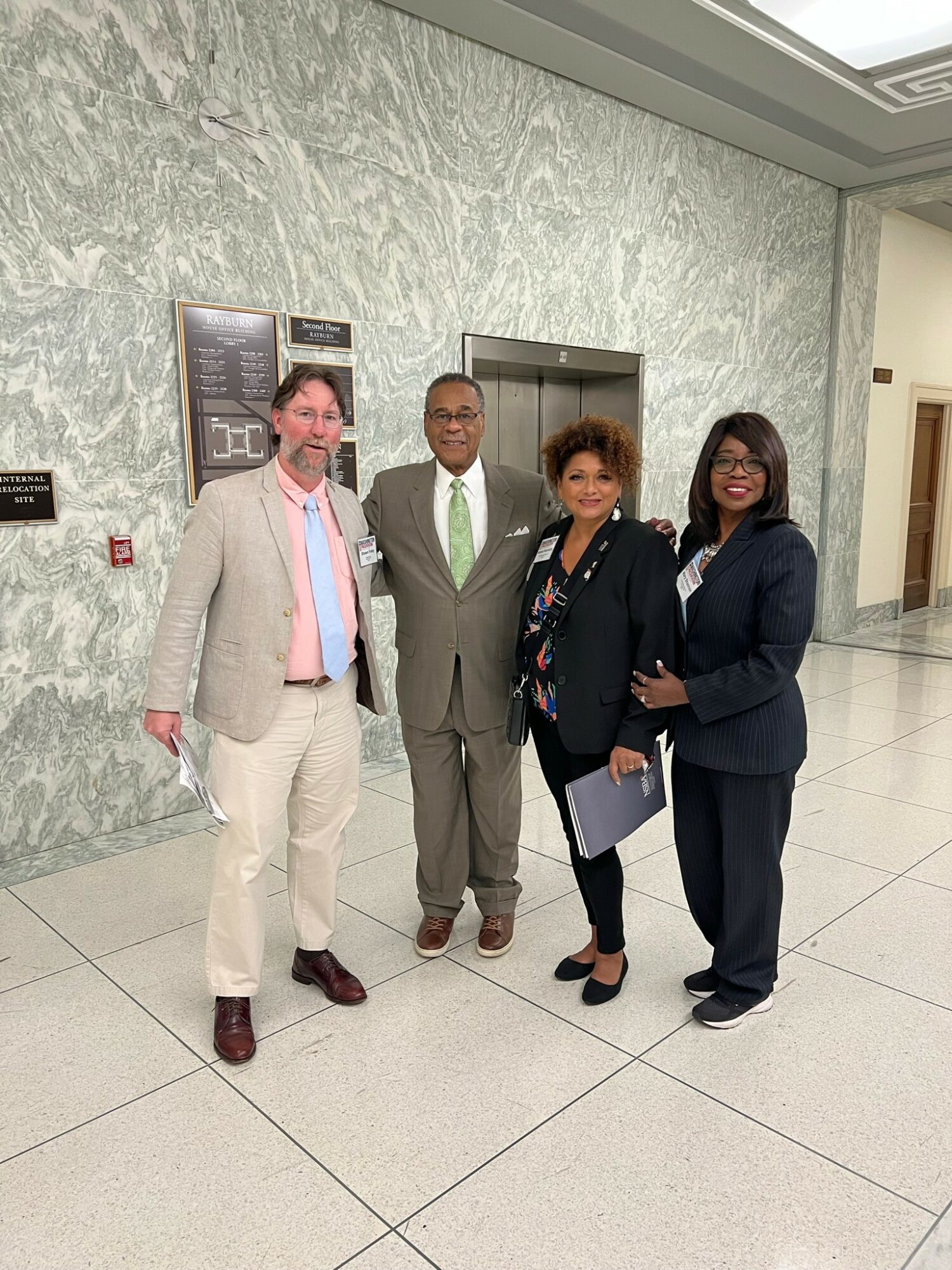 Four people standing together in a hallway with a marble wall and elevator behind them.
