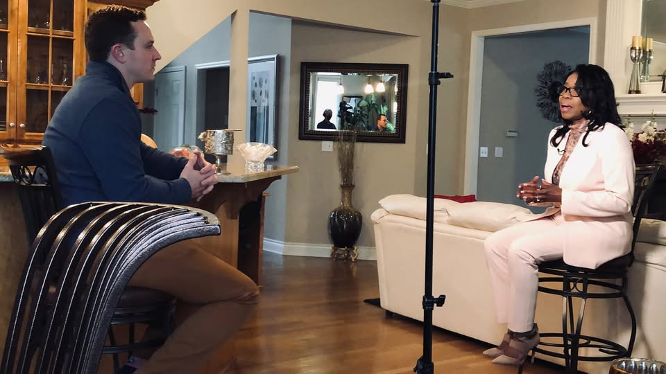 Two people sit facing each other in a living room, engaged in conversation, with a camera setup nearby.