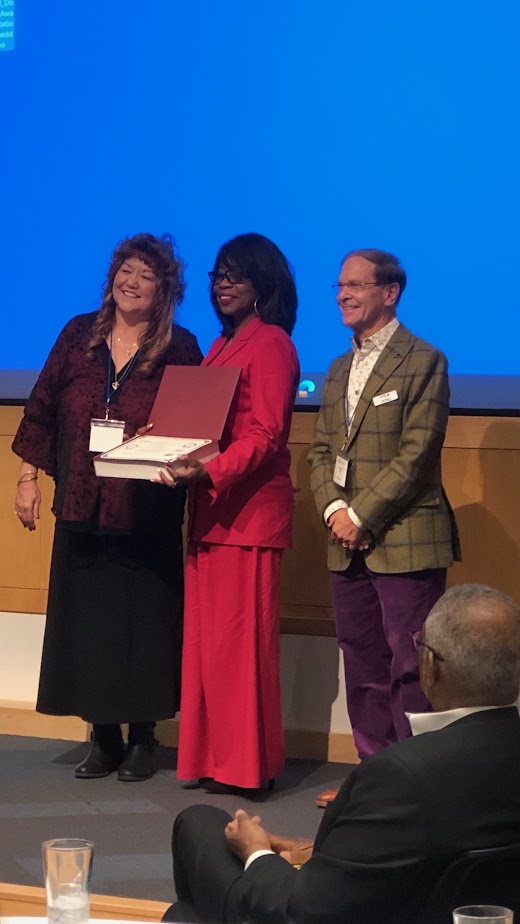 Three people on stage, one holding a certificate, with a blue background and an audience member in foreground.
