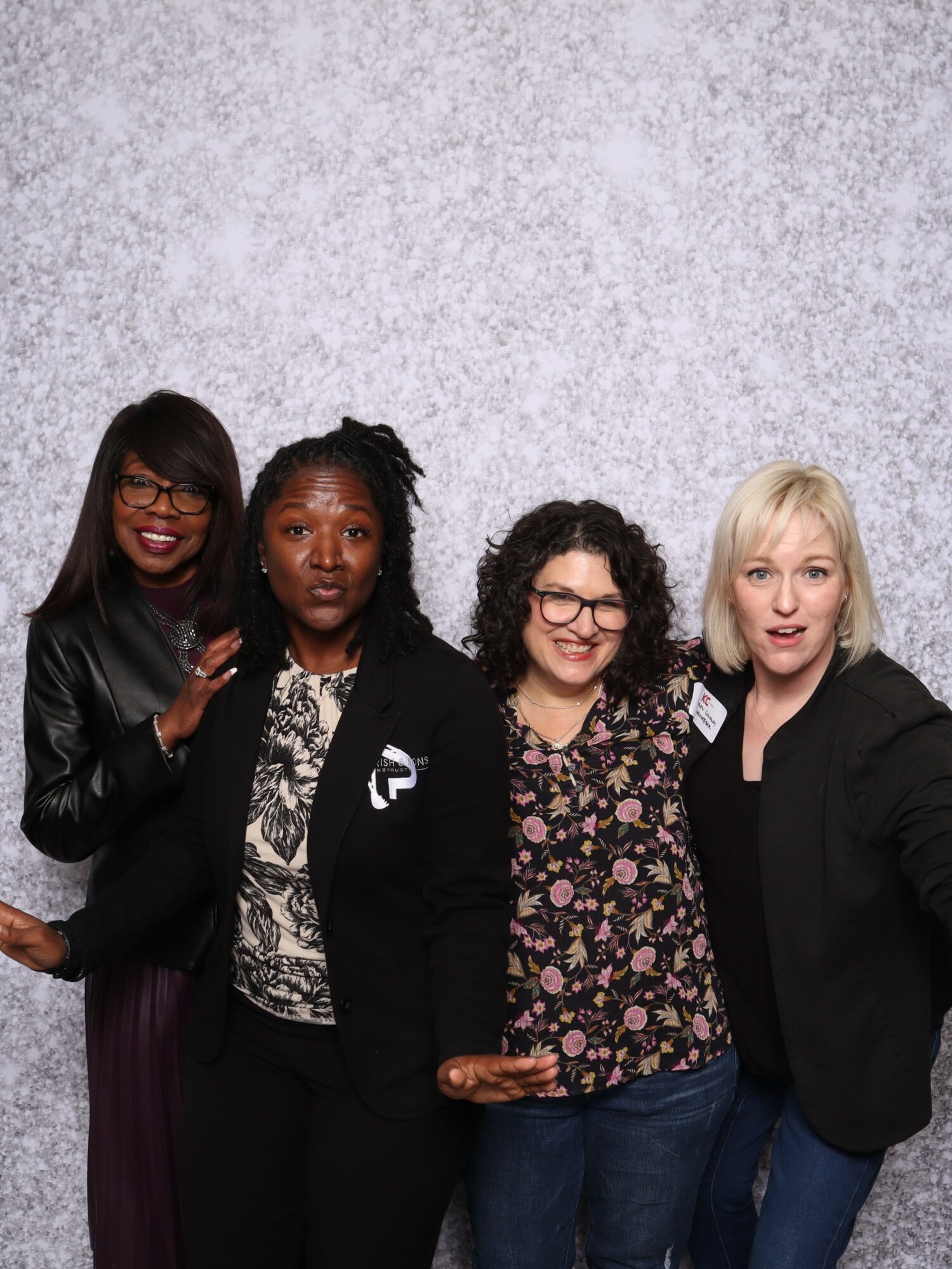 Four women standing together, smiling, in front of a light textured background.