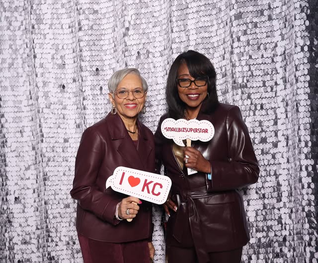 Two women standing in front of a silver sequin backdrop, holding signs, smiling, wearing dark leather jackets.