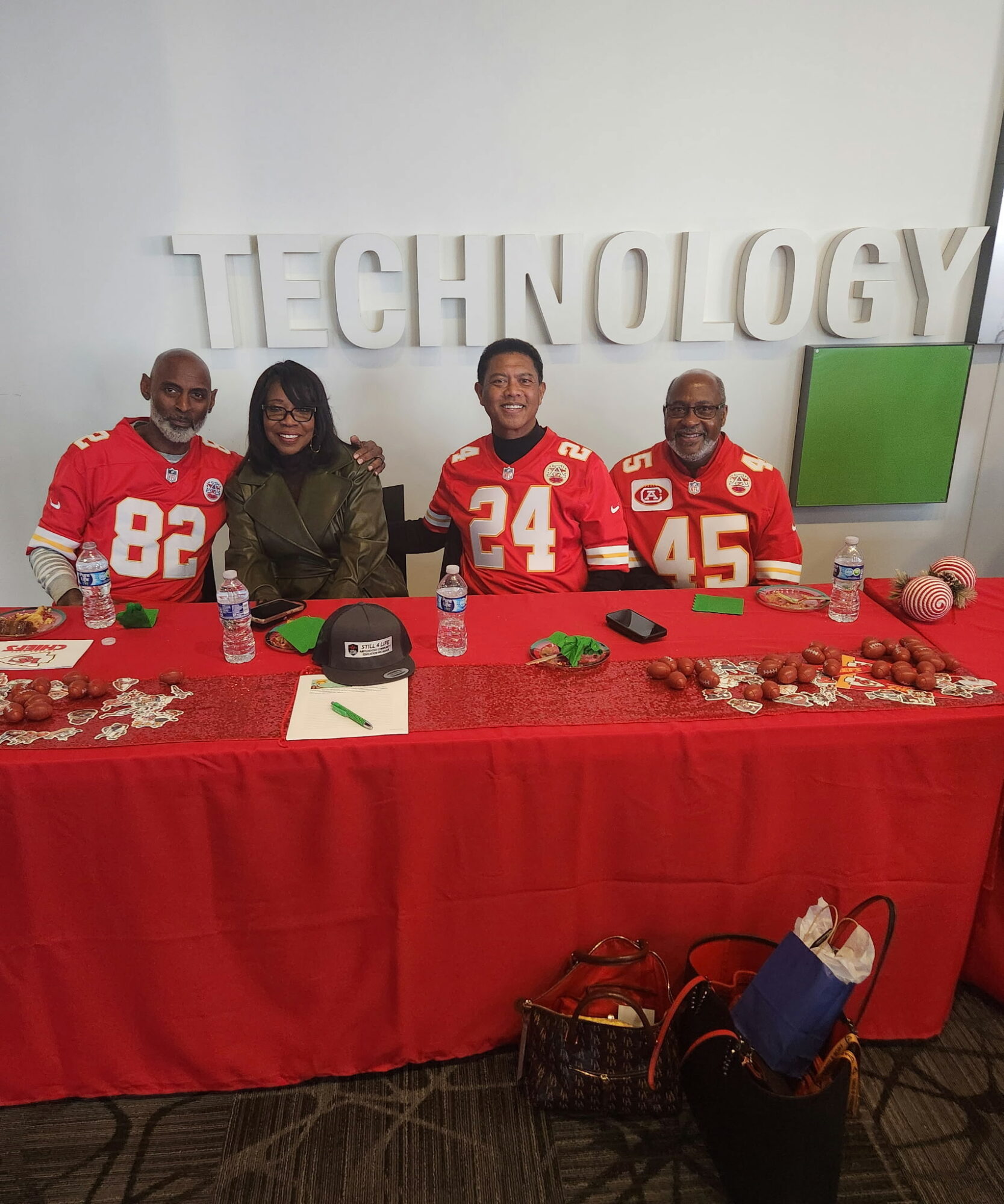 Four people sit at a table with a red tablecloth, in front of a wall with large white letters spelling 'TECHNOLOGY'.