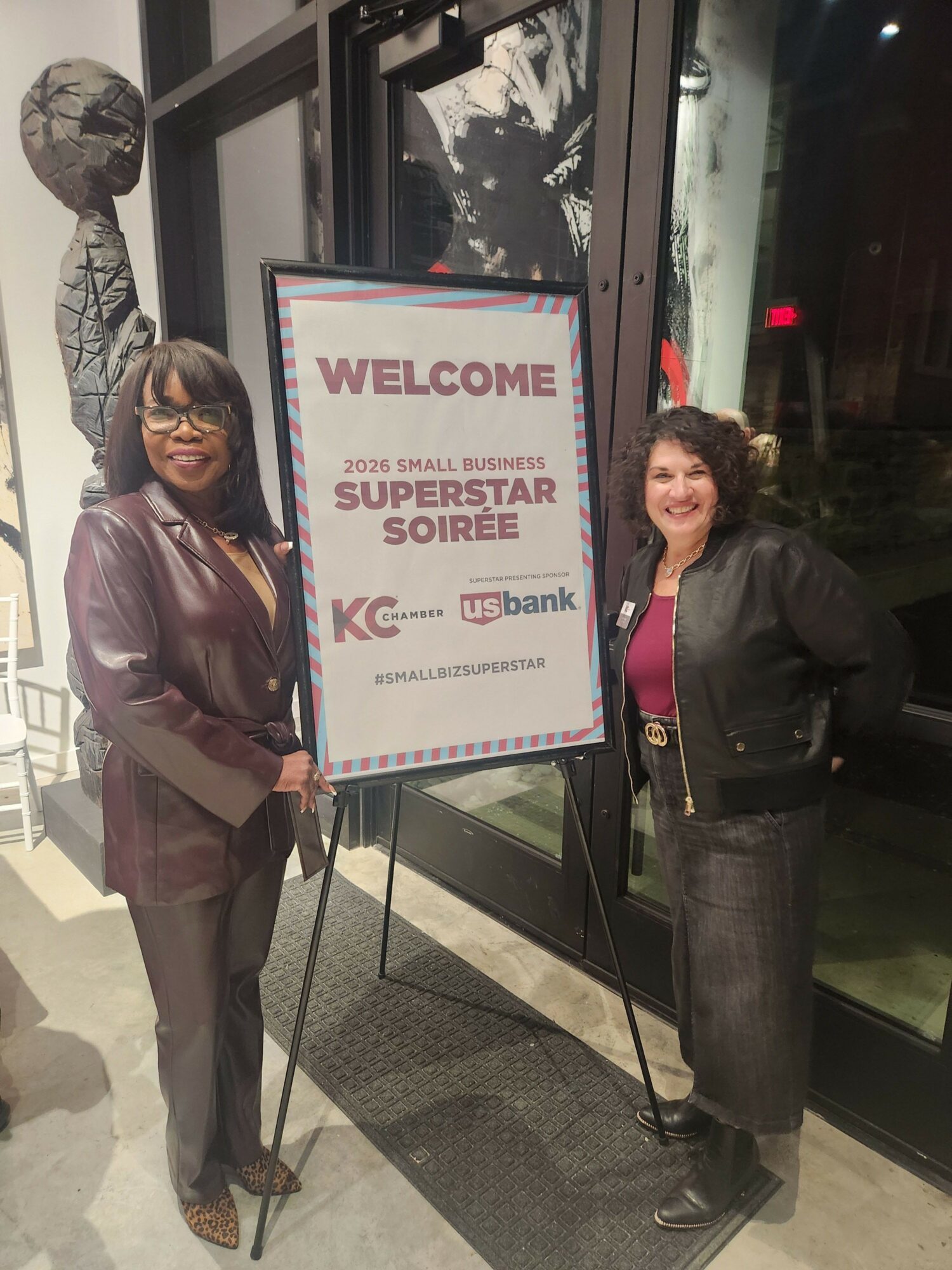 Two women standing outside a building near a sign welcoming to a sports event, with one woman holding a purse.