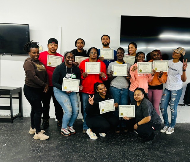 Group of people holding certificates, standing in a room with a large screen and a TV, smiling and posing for a photo.