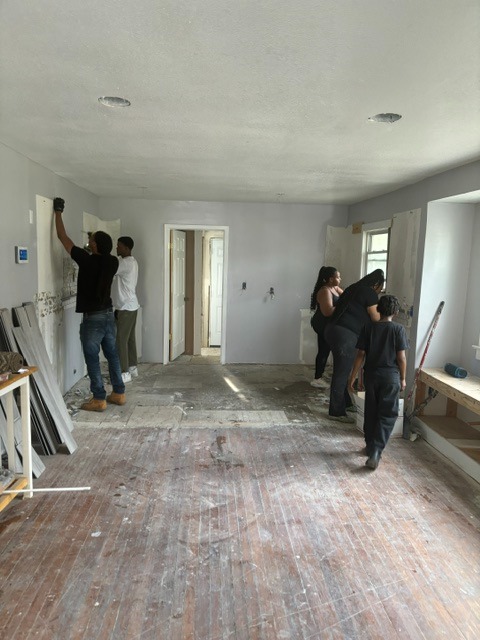 Group of people working on a room with white walls and large windows, some painting the ceiling, others observing or working.