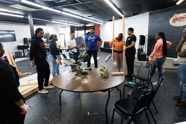 Group of people standing in a room with a table and chairs, some looking at a person in a blue shirt, in a workshop or meeting space.