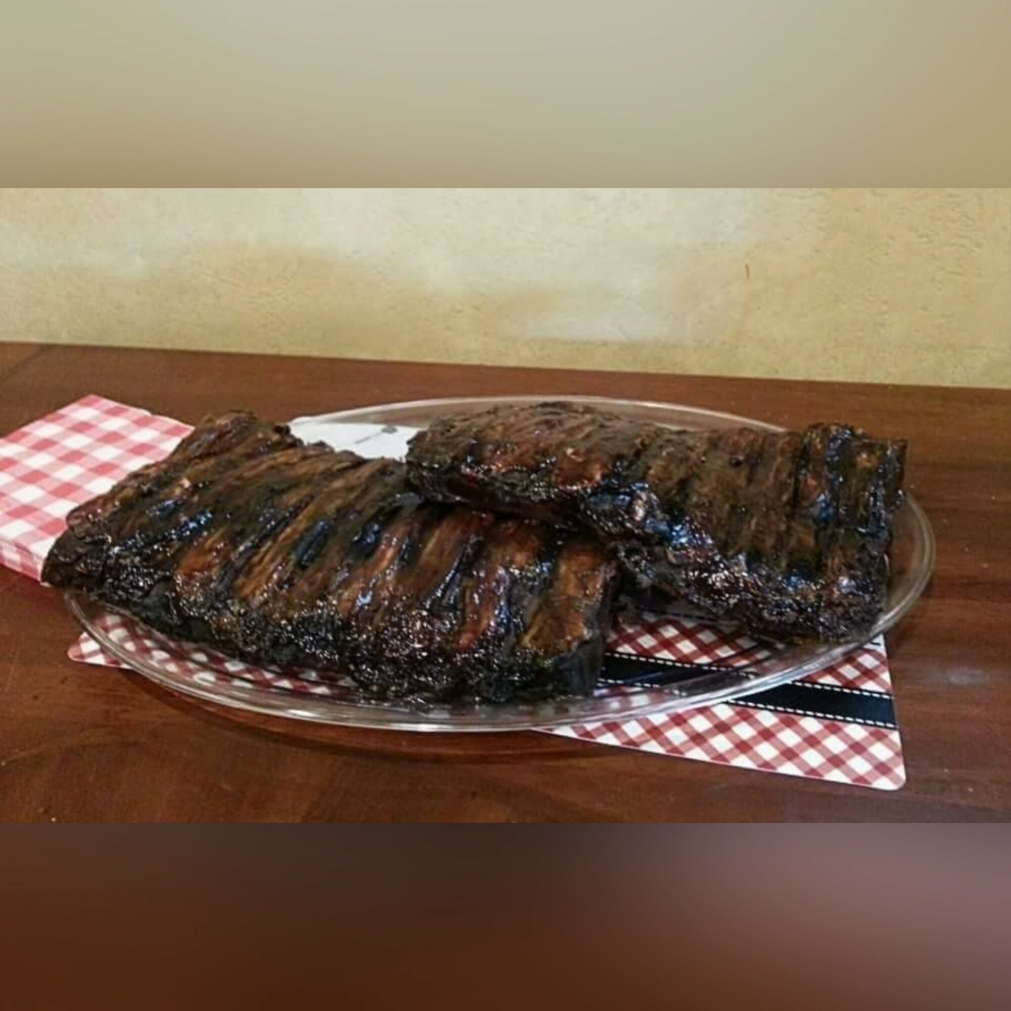 Two grilled ribs on a plate with a checkered napkin, on a wooden table, against a beige wall.