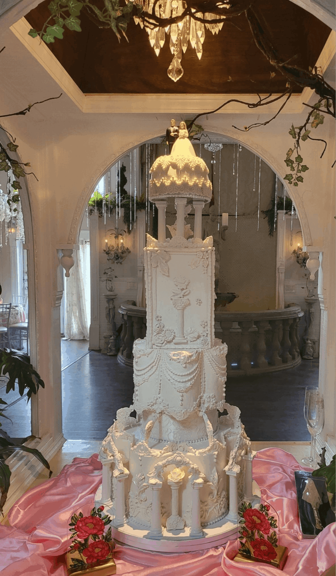 Elegant multi-tiered white wedding cake with intricate designs, decorated with pink flowers, in a decorated venue with arches and chandelier.