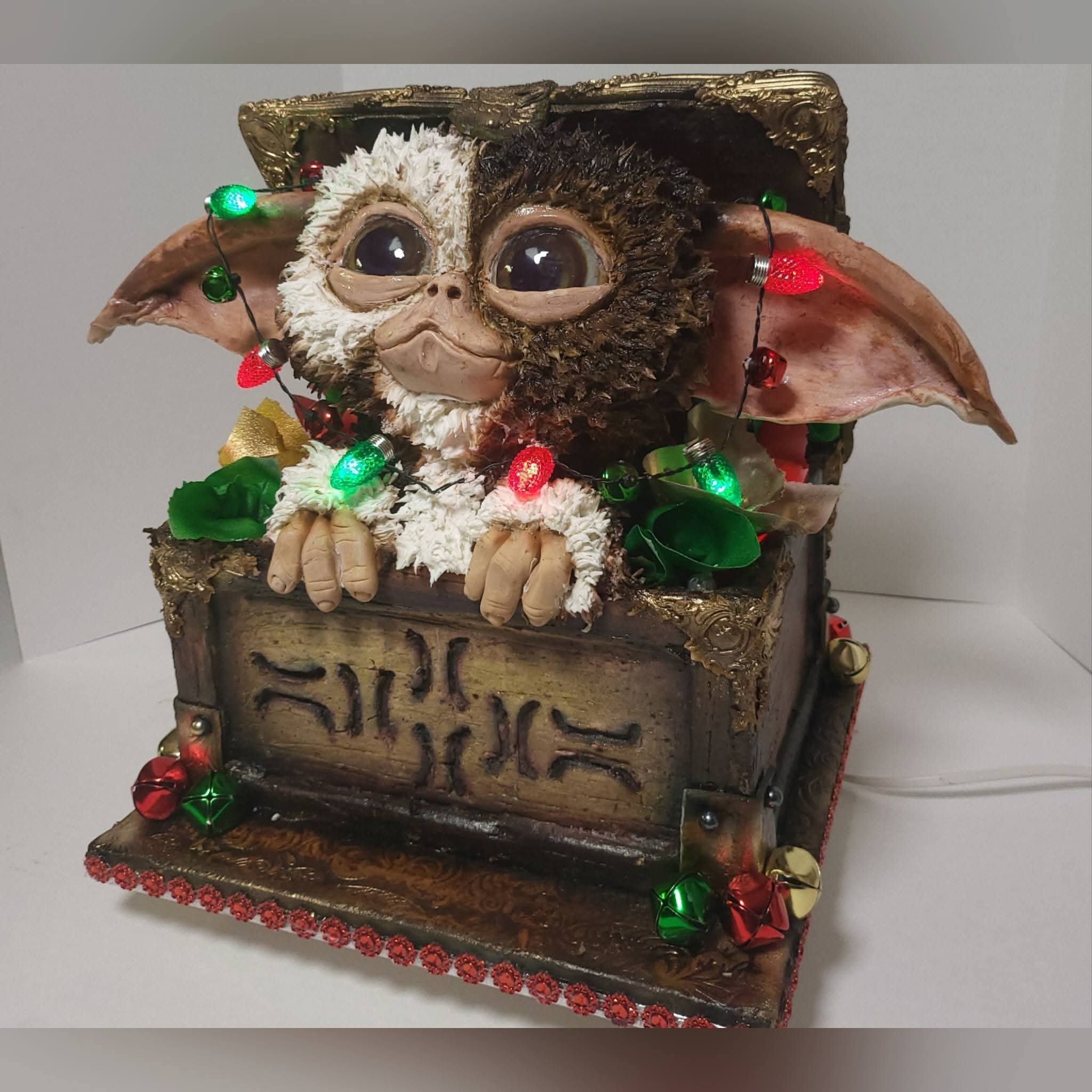 Decorative creature with large eyes, furry face, and bat-like ears, inside a decorated box with lights and ornaments.
