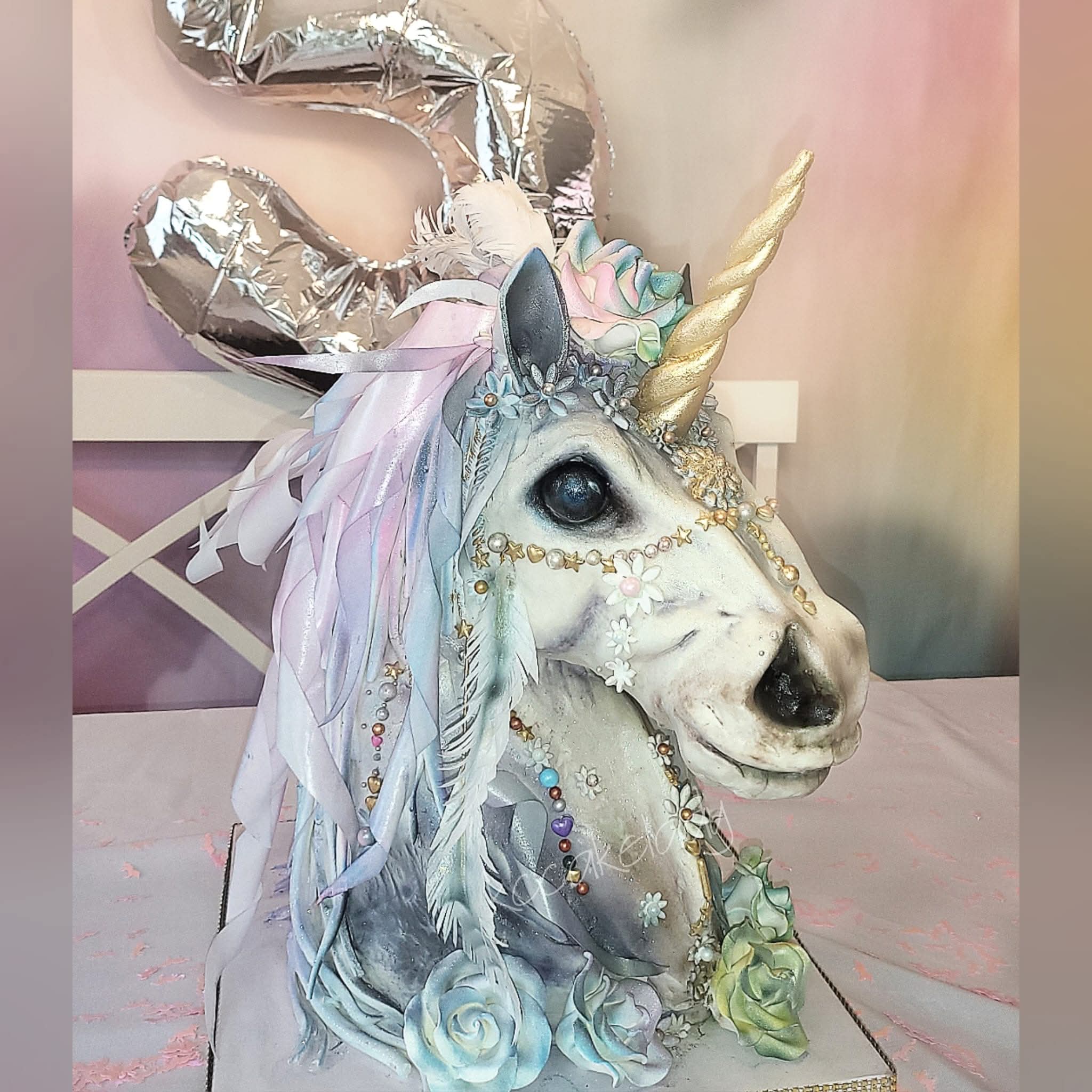 Decorated unicorn cake with balloons, flowers, and pastel colors, featuring a golden horn and detailed face.