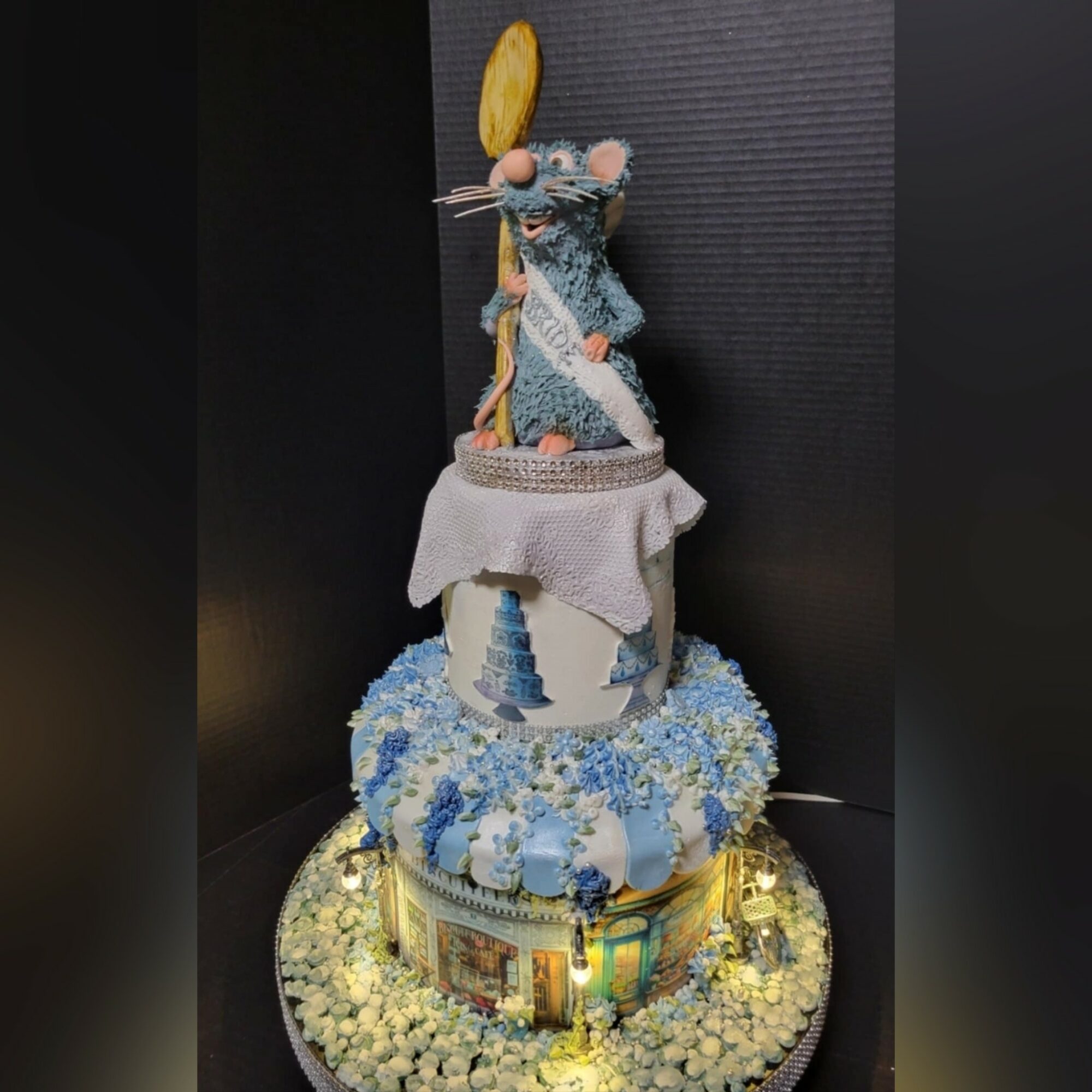 Decorative cake with a mouse figurine holding a spoon and a cookie, surrounded by flowers and a building image, on a black background.