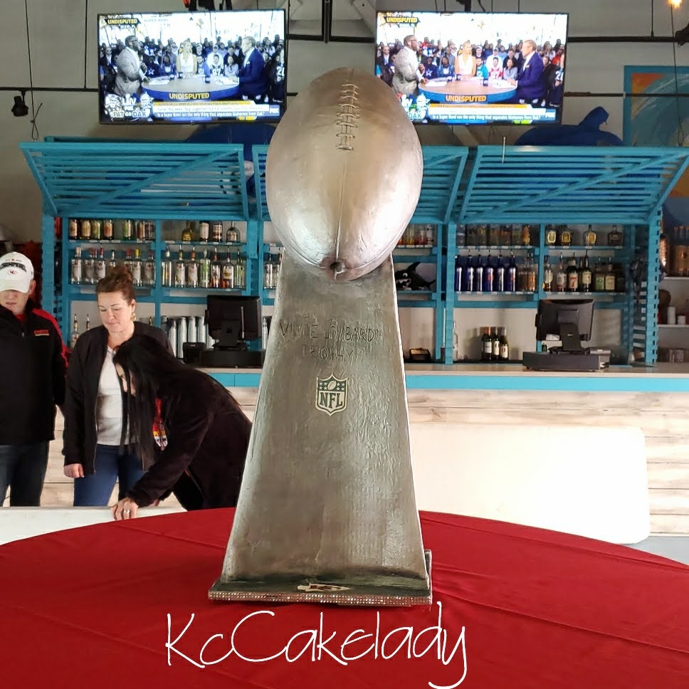 Large football trophy displayed on a red cloth with people and a bar in the background.