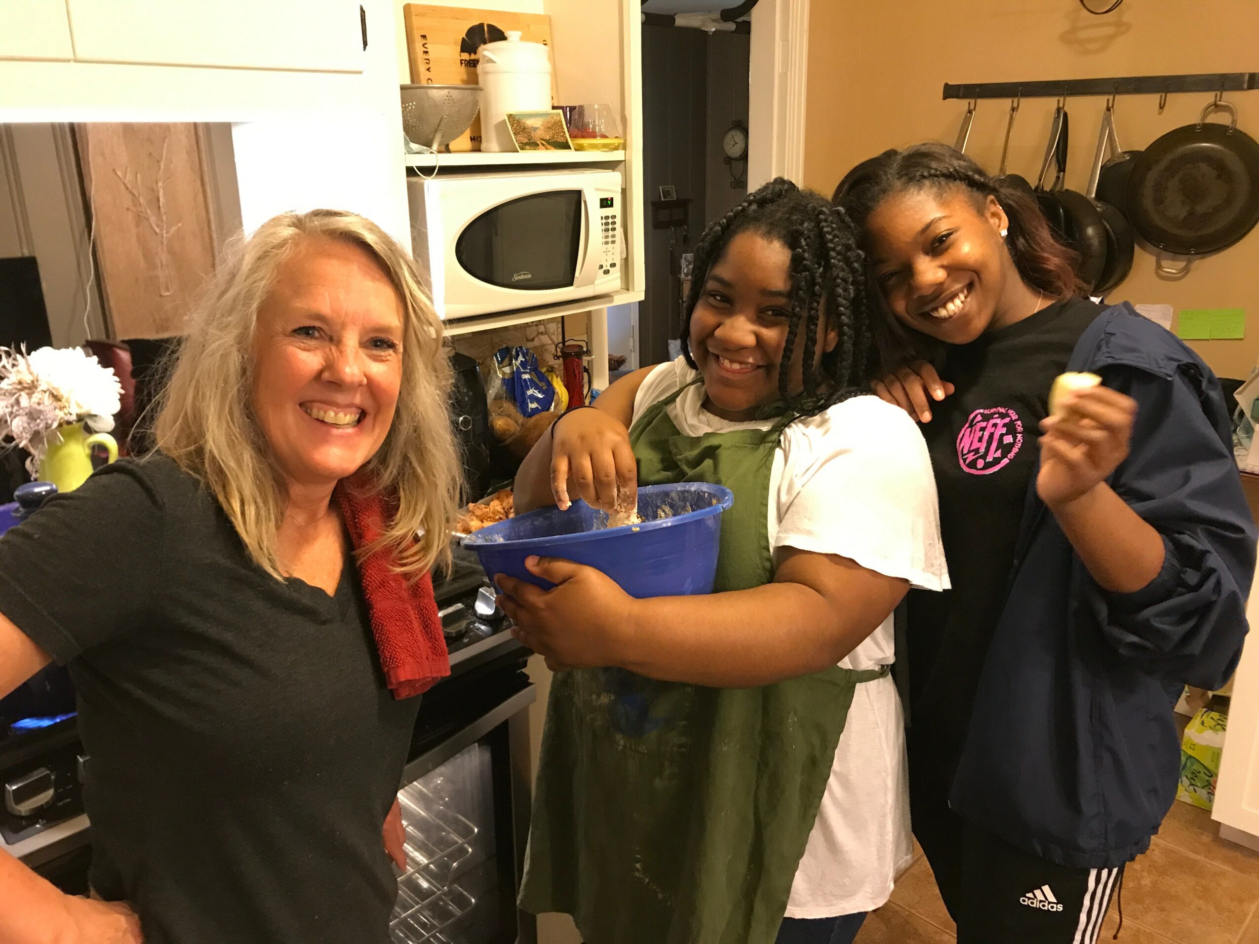 Three smiling people in a kitchen, two children and an adult woman, standing close together, with kitchen appliances in the background.