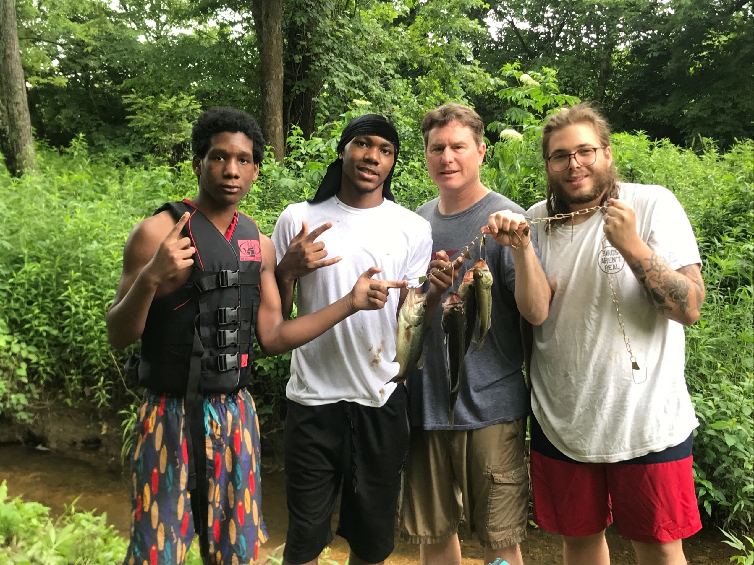 Four people standing outdoors in a green wooded area, holding fish and making gestures, smiling at the camera.