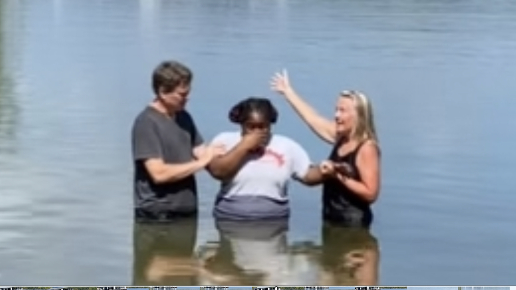 Three people standing in water, one woman raising her hand, another woman and a man holding hands, smiling.