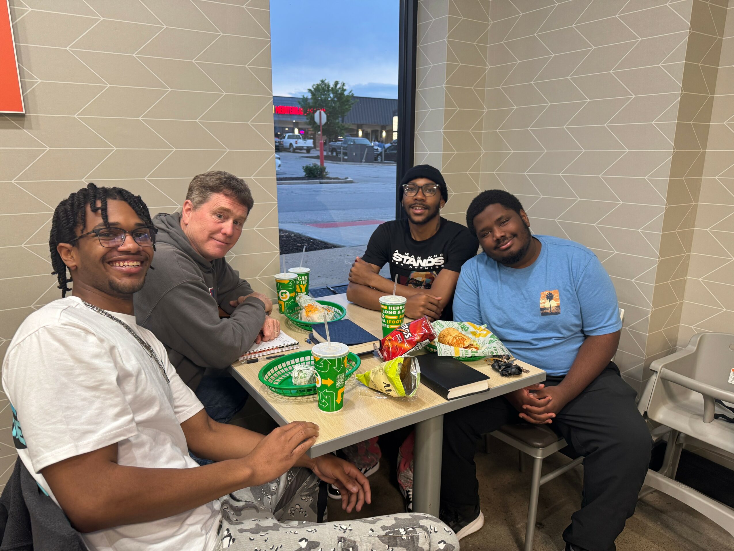 Four people sitting at a table inside a fast-food restaurant, smiling, with food and drinks on the table.