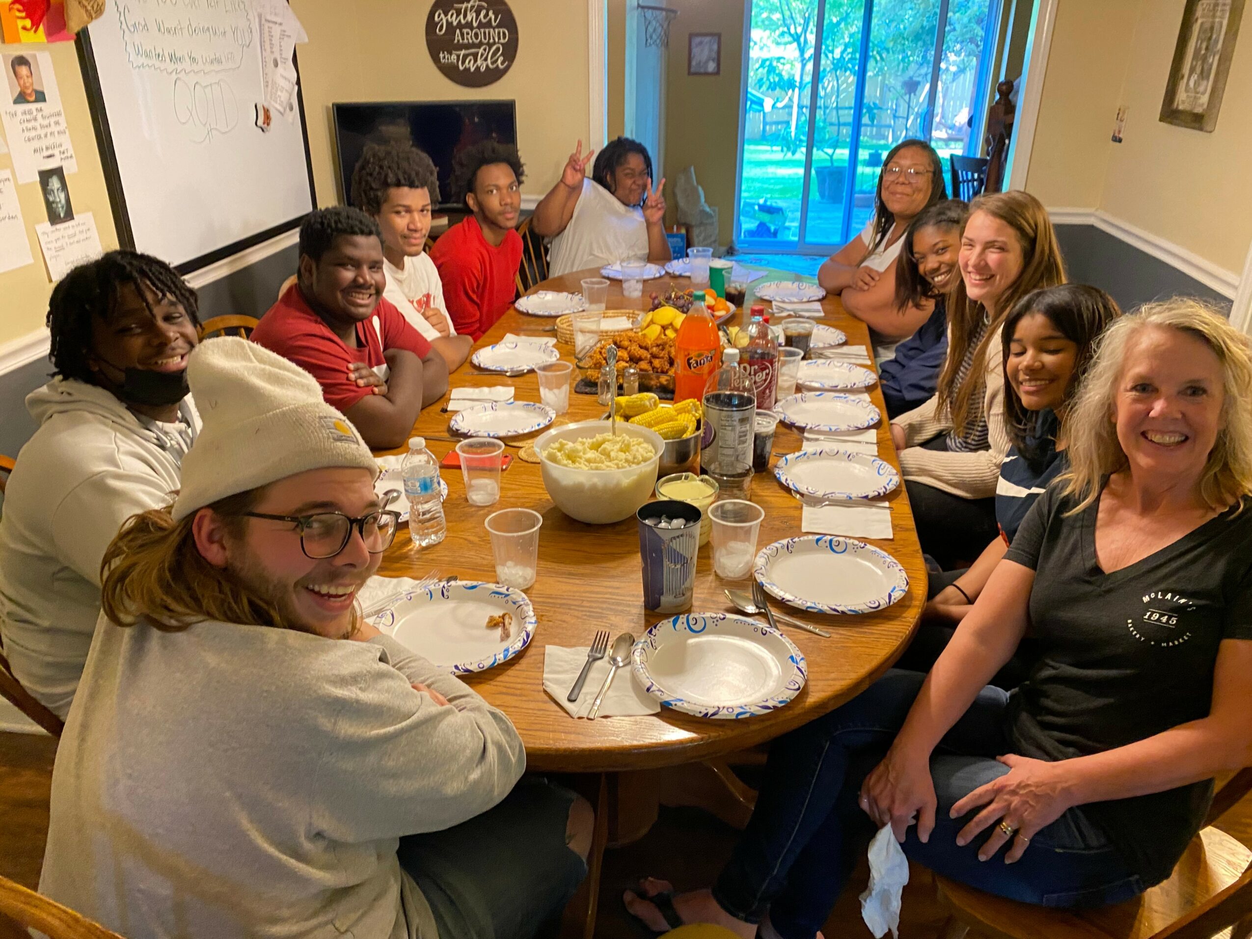 Group of people sitting around a dining table with food and drinks, smiling, in a room with a window showing trees outside.