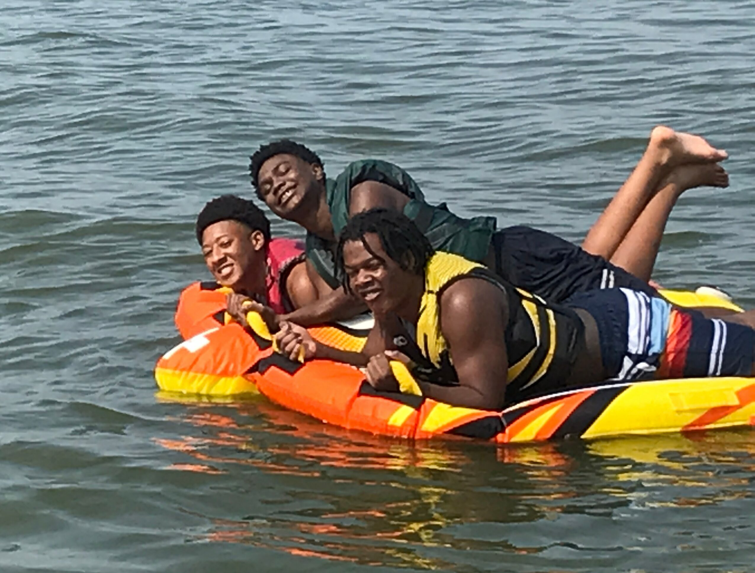 Three people lying on a yellow and orange inflatable raft in the water, smiling and enjoying a day at the beach.