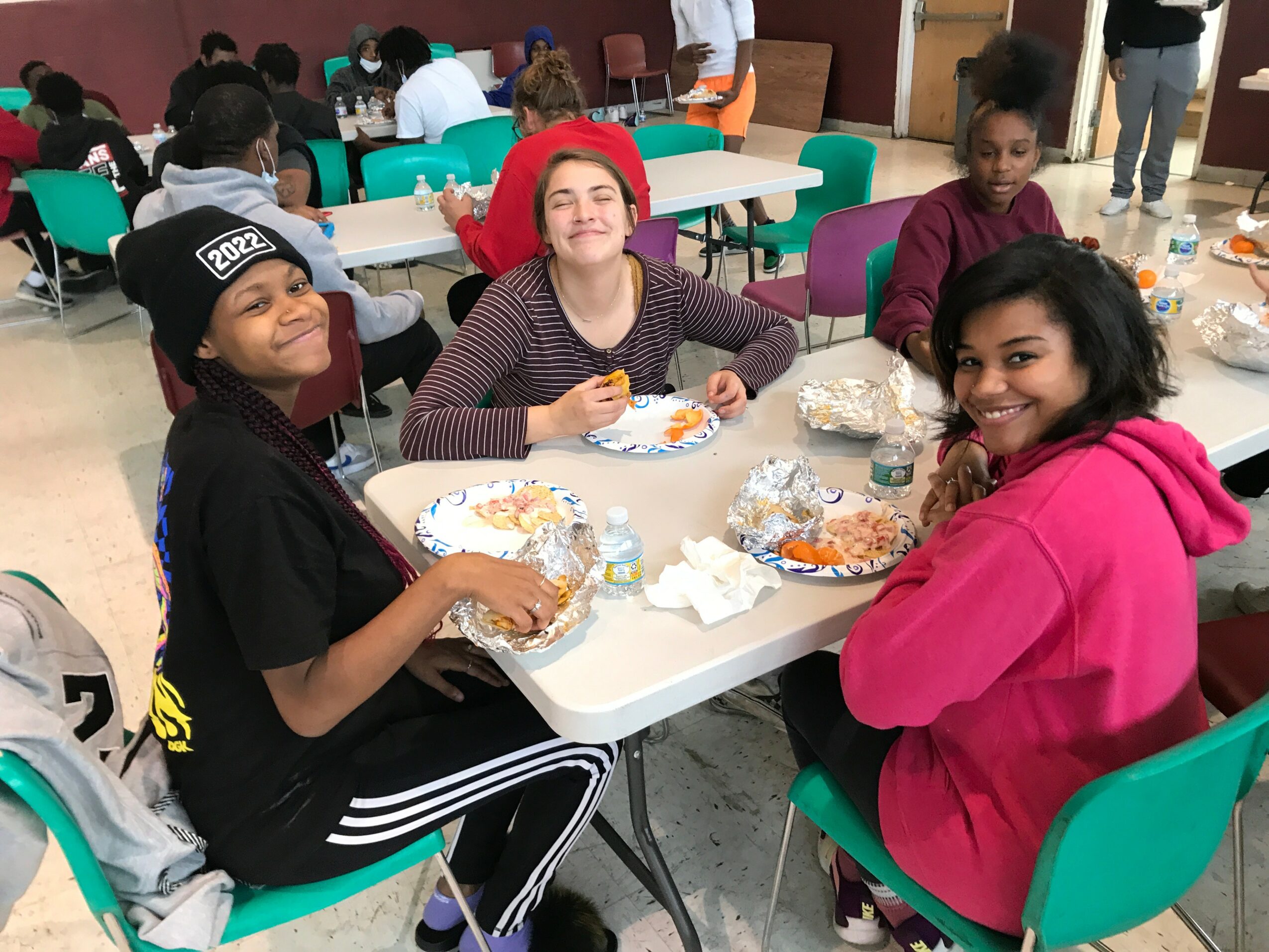 Four children sit at a table eating food, smiling, in a room with other people and tables in the background.