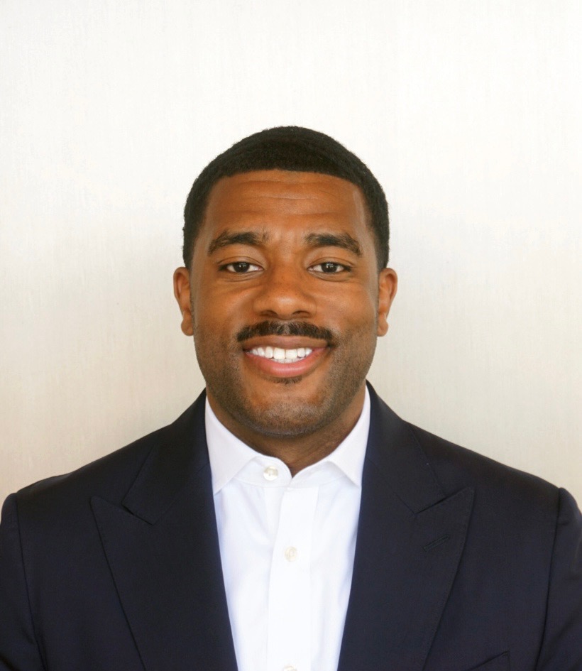 A man in a dark suit and white shirt smiling against a plain background.