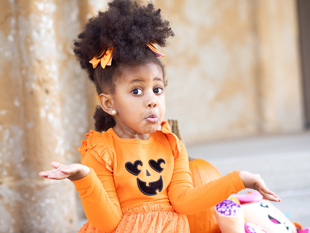 Young girl with curly hair and orange dress sitting outdoors, holding toys, with a surprised expression.