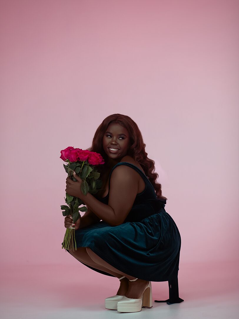 Woman with long hair holding a bouquet of pink roses, squatting on a pink background, smiling.