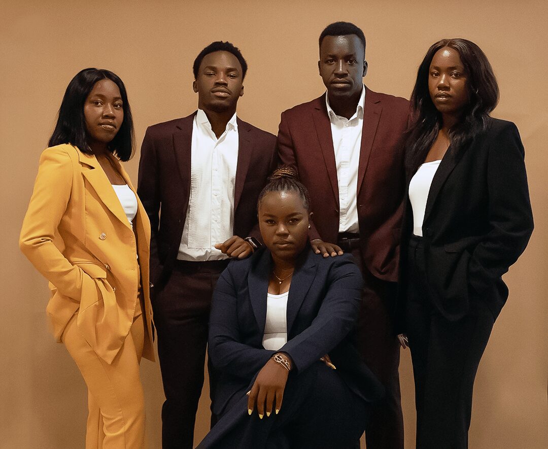 Group of five people standing together, four women and one man, dressed in formal attire, against a plain background.
