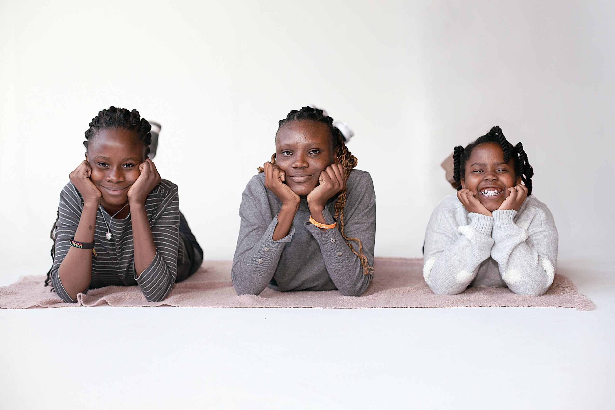Three children lying on their stomachs with hands on cheeks, smiling, against a plain white background.