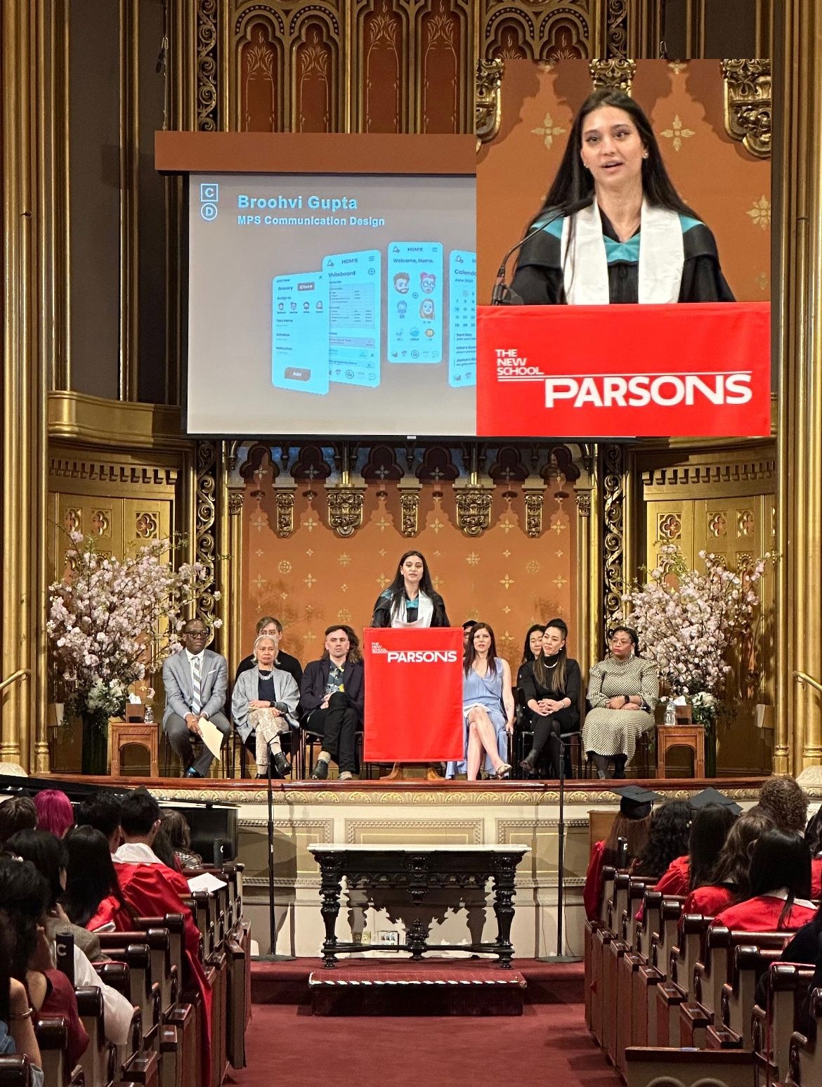 Woman speaking at a podium on stage with seated panel and audience, large screen behind displaying presentation slides, ornate decor.