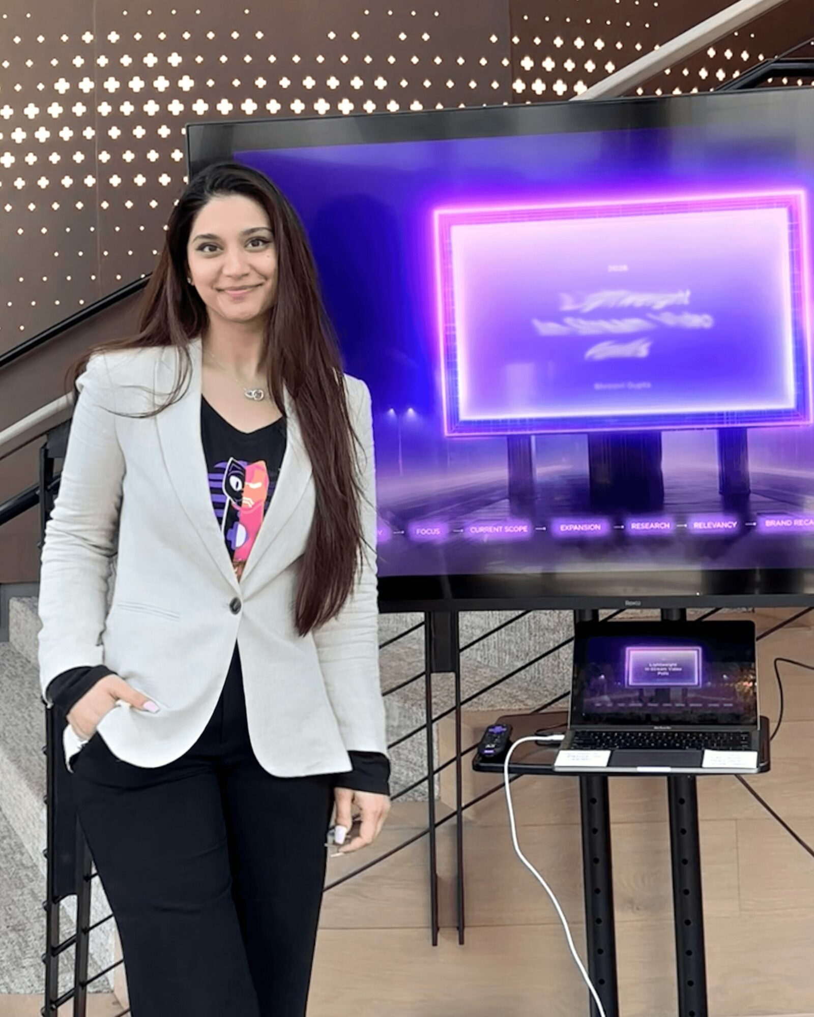 Woman in white blazer and black pants standing next to a large screen and laptop, smiling indoors.