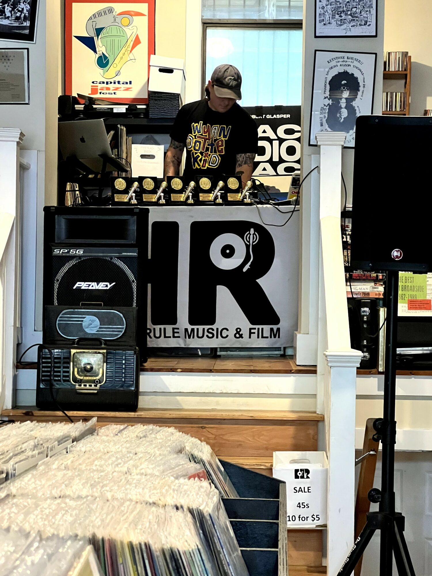 Person wearing a cap and black shirt behind a DJ setup with a large logo reading 'R' and 'RULE MUSIC & FILM' in a room with posters and equipment.