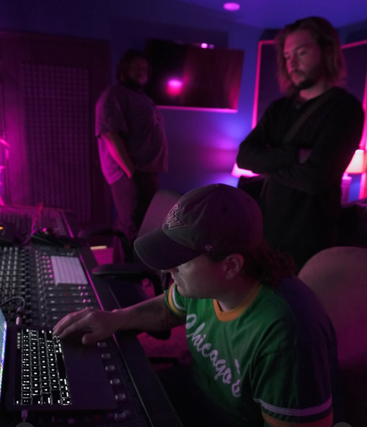 Person wearing a cap and green shirt working on audio equipment, with two people in the background in a dimly lit room with purple lighting.