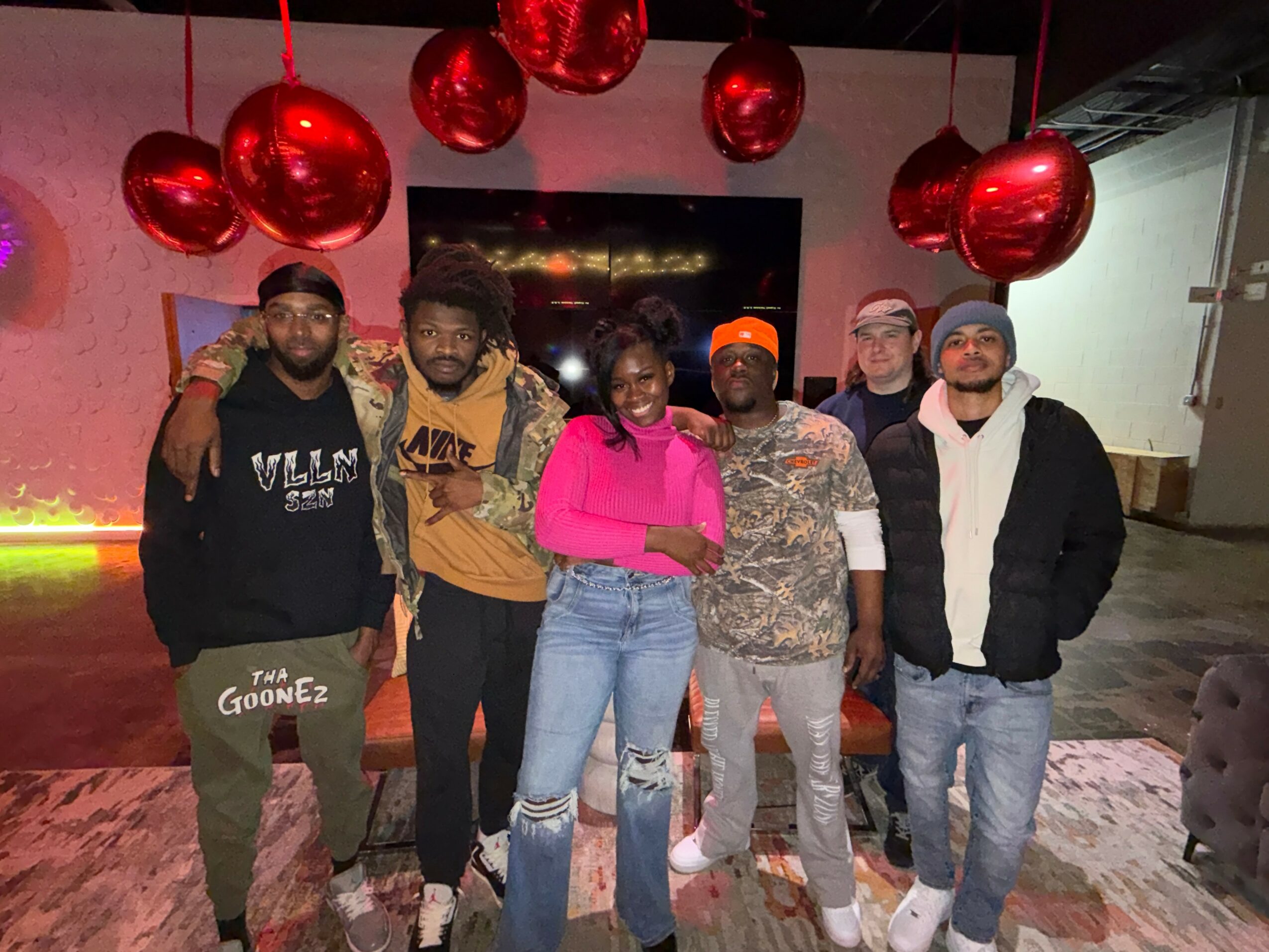 Group of six people standing indoors with red hanging decorations, smiling, some with arms around each other.