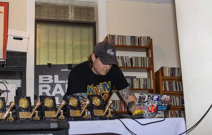 Person wearing a cap and glasses, working at a DJ setup with trophies and books in the background.