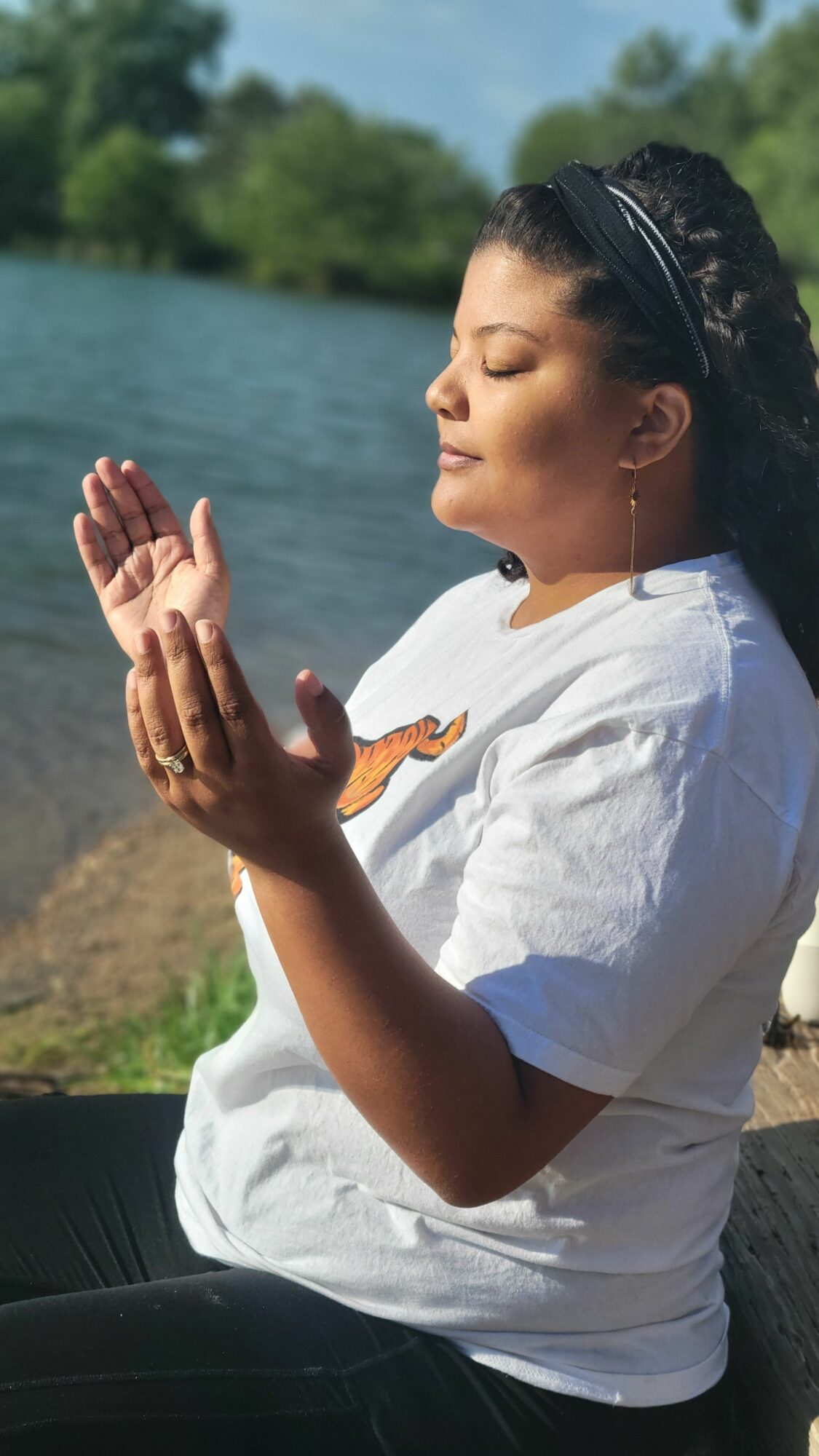 Person practicing yoga outdoors near water, with trees in the background, wearing a white shirt and black pants.