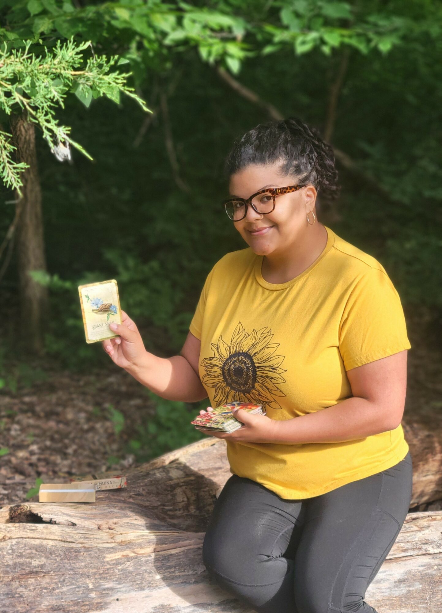 Smiling woman with glasses in a yellow shirt holding a small book outdoors, surrounded by green trees.