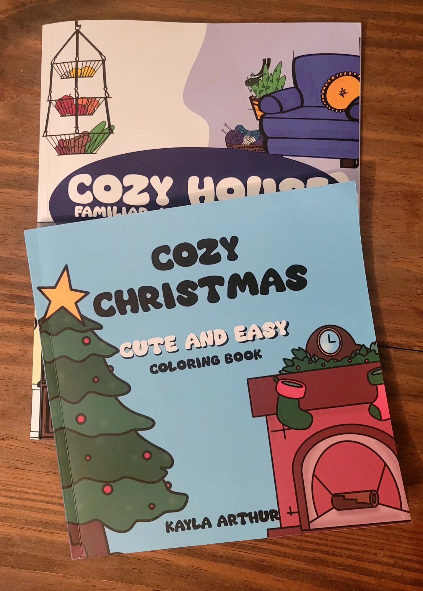 Two children's coloring books on a wooden surface, one titled 'Cozy Christmas' and the other 'Cozy Home,' with festive illustrations.