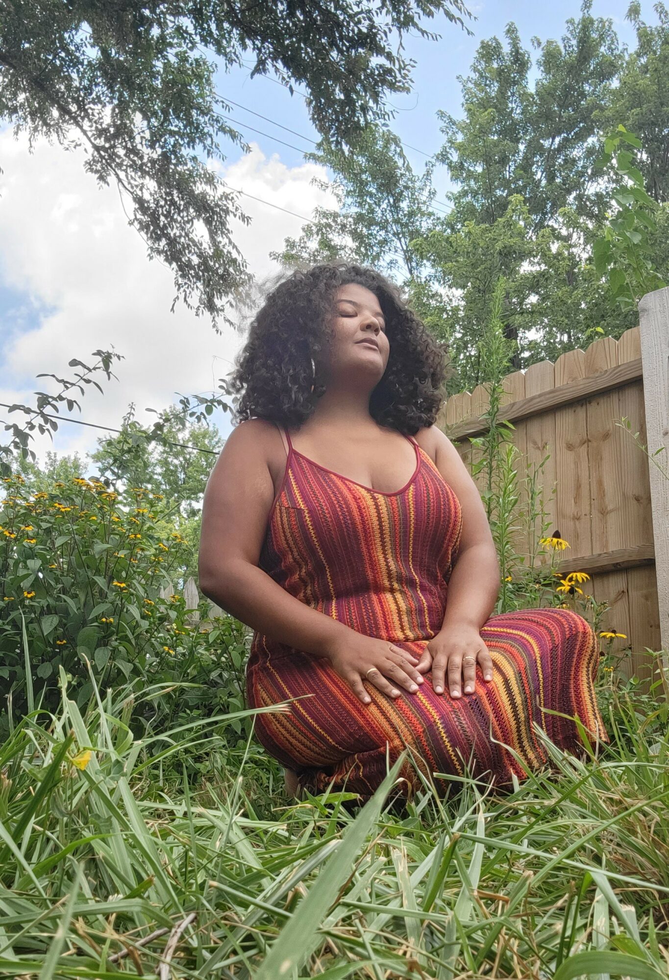 Woman with curly hair sitting outdoors on grass, wearing a striped dress, with trees and a wooden fence in background.
