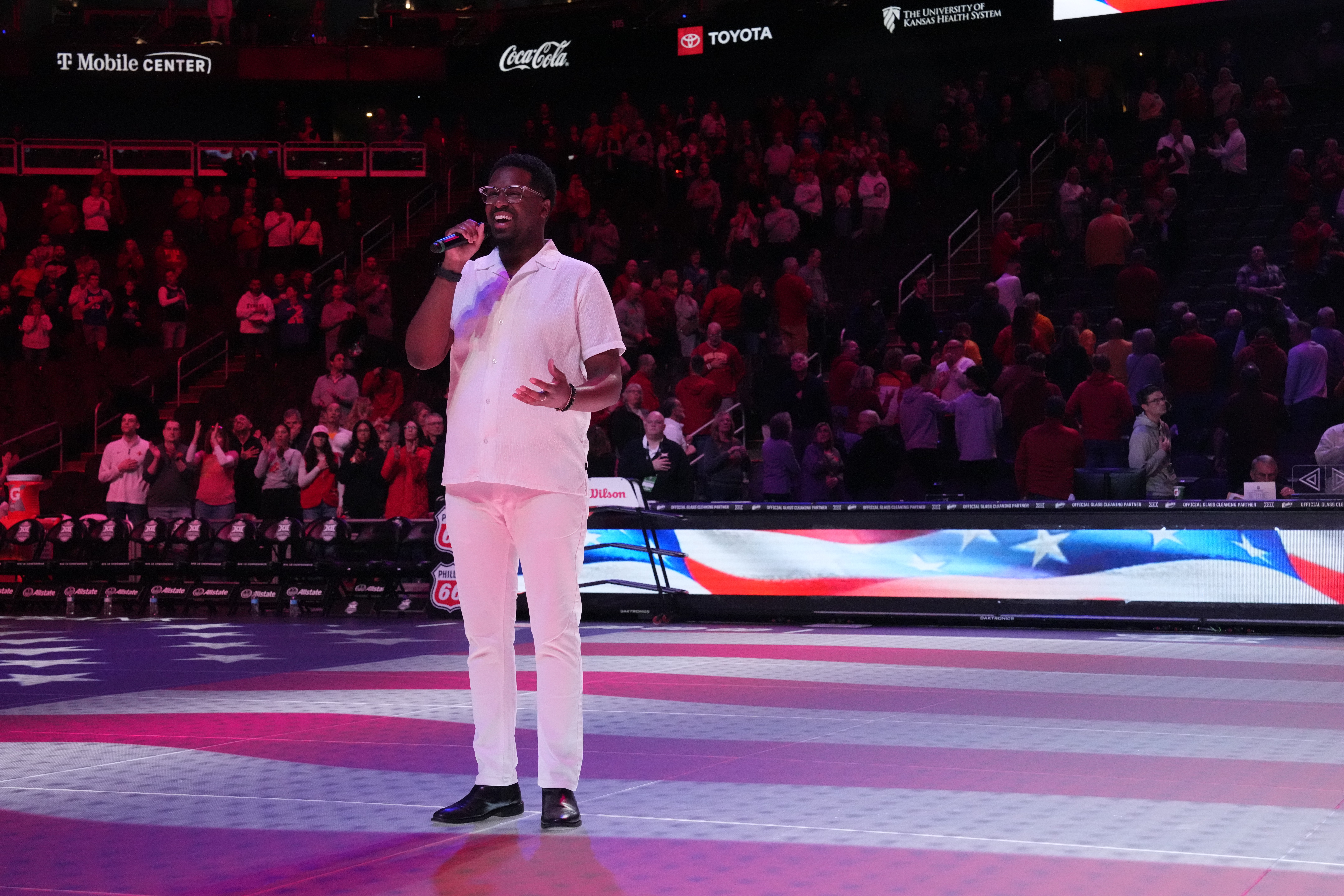 Person in white outfit singing on stage with audience in background, American flag design on floor, red lighting.