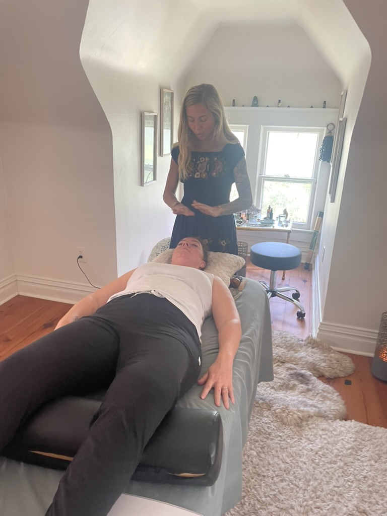 A woman lies on a massage table with her eyes closed, while a girl stands nearby in a room with a window and a stool.