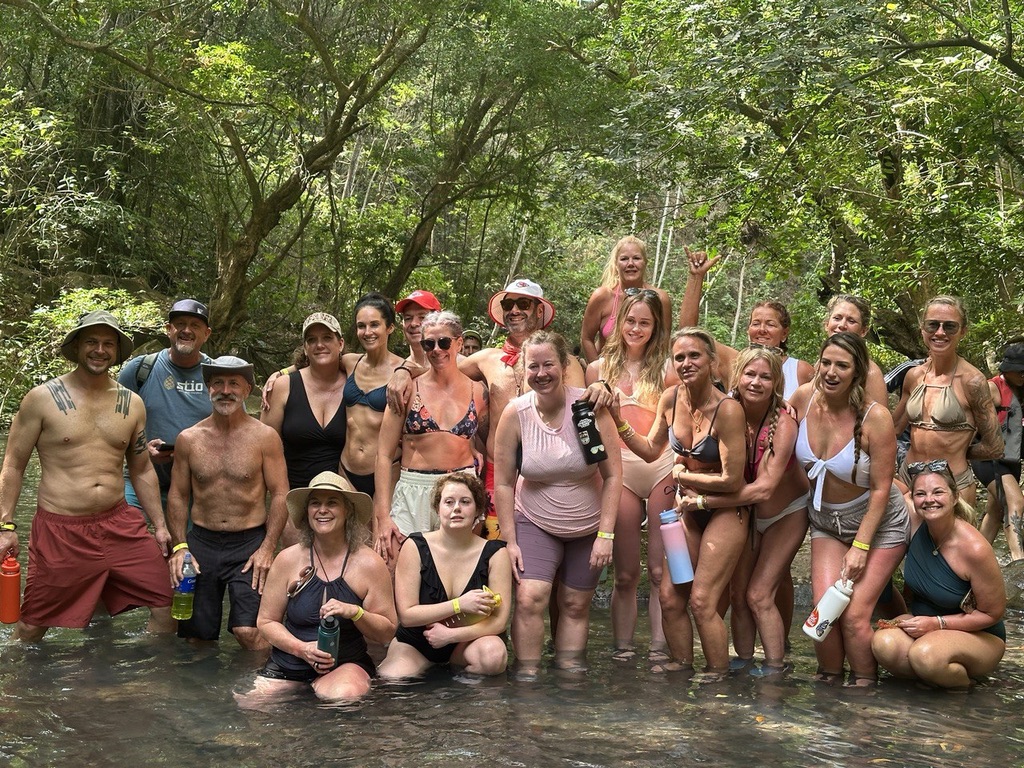Group of people standing in water outdoors surrounded by trees, some wearing swimsuits and hats, smiling and posing for the photo.