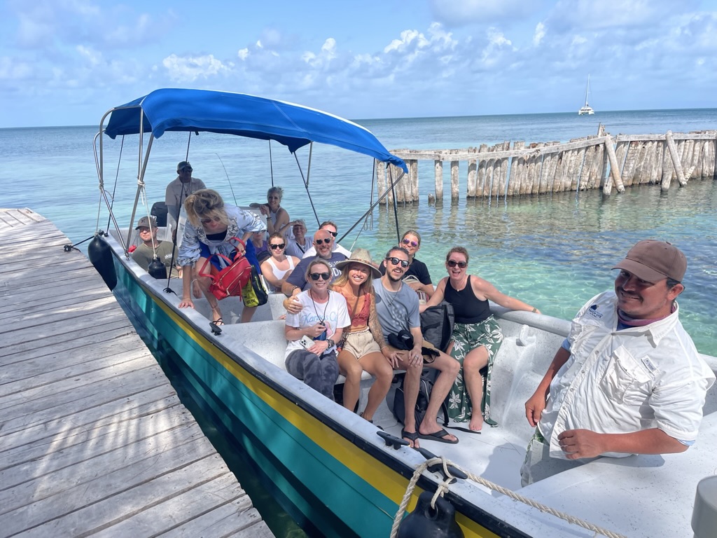 Group of people sitting on a boat docked at a pier, with a man standing at the front, ocean and sky in background.