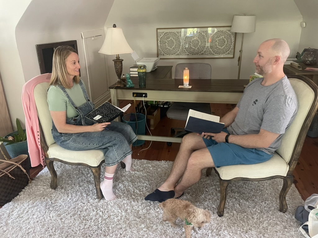 Two people sit in chairs facing each other in a room, with a small dog on the floor between them.