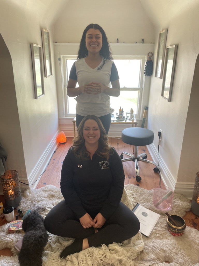 Two women sitting and standing in a hallway with a window behind them, smiling, surrounded by various objects and decorations.