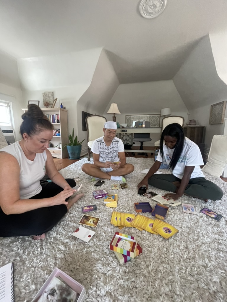 Three women sit on a carpeted floor playing a card game, with a bookshelf and decorative items in the background.