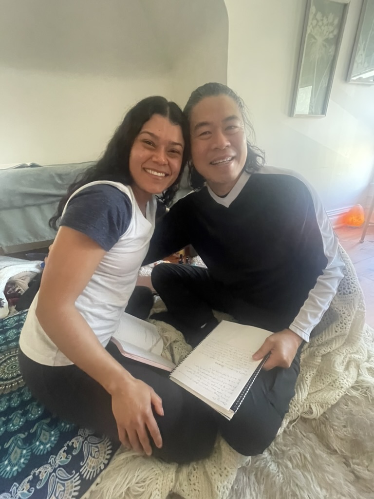 Two smiling women sitting on a bed with notebooks, close together indoors, with a window in the background.