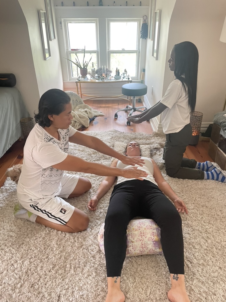 Two women are performing a massage or therapy on a person lying on a cushion in a living room with a window and plants in the background.