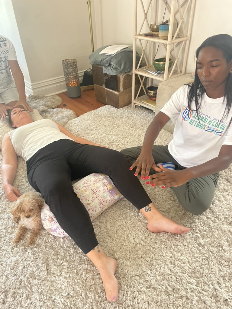 Person lying on a rug with a dog nearby, another person kneeling beside them touching their hand, in a cozy room.