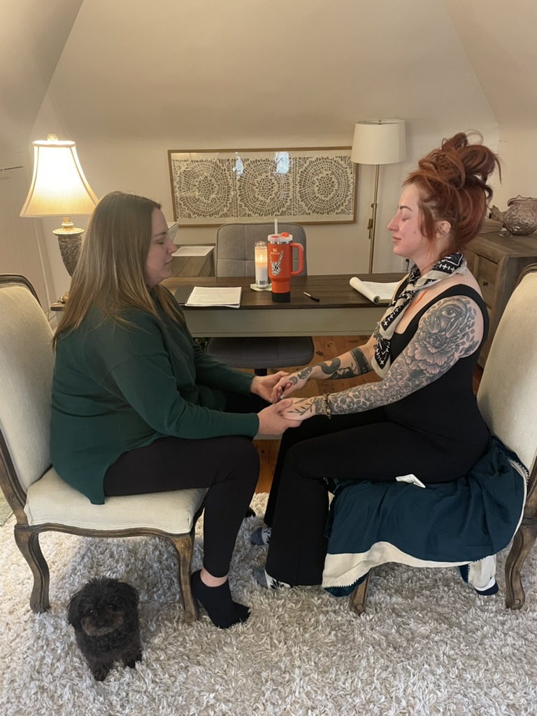 Two women sit facing each other holding hands in a living room, with a small black dog on the carpet nearby.