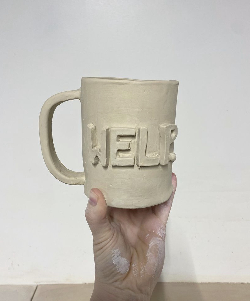 Hand holding a beige ceramic mug with the word 'HELLO' embossed on it, against a plain white background.